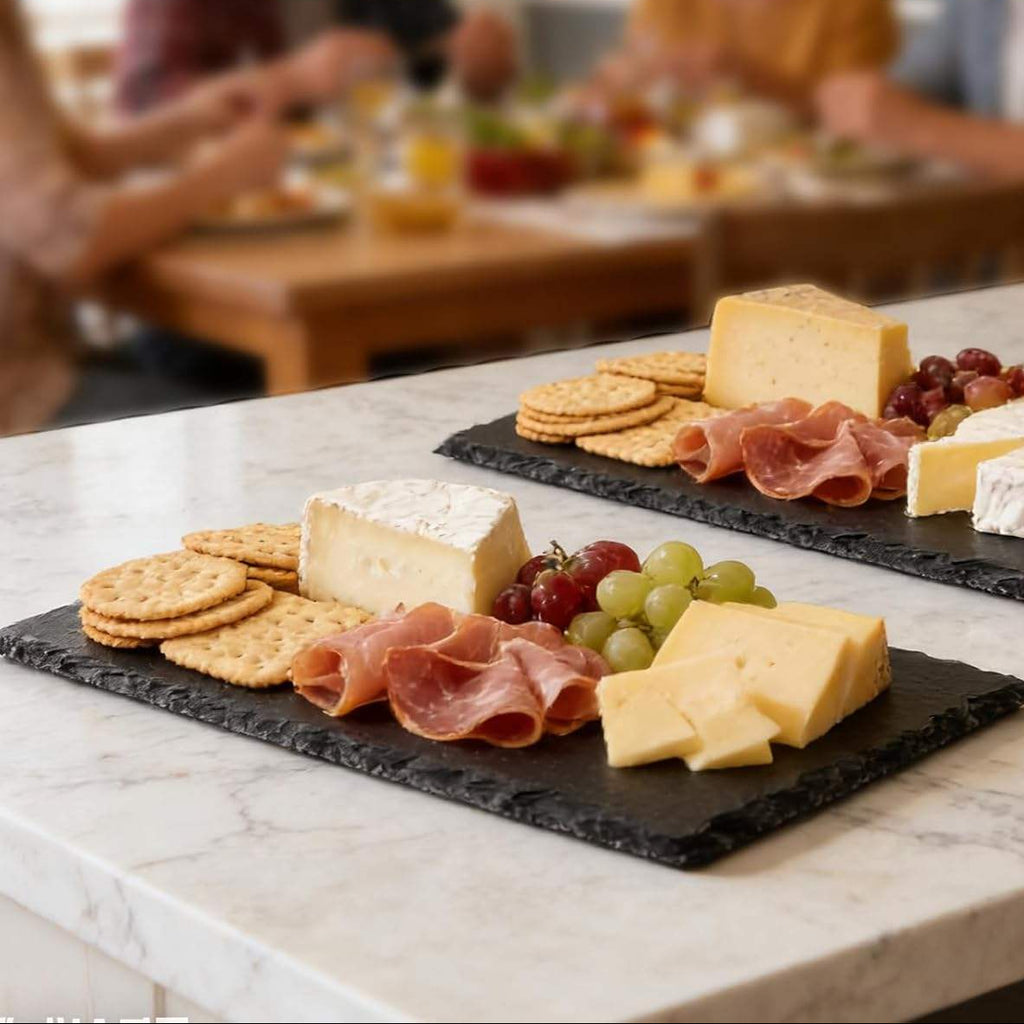 Plates of cheese, crackers, and grapes on a slate plate with people in the background.